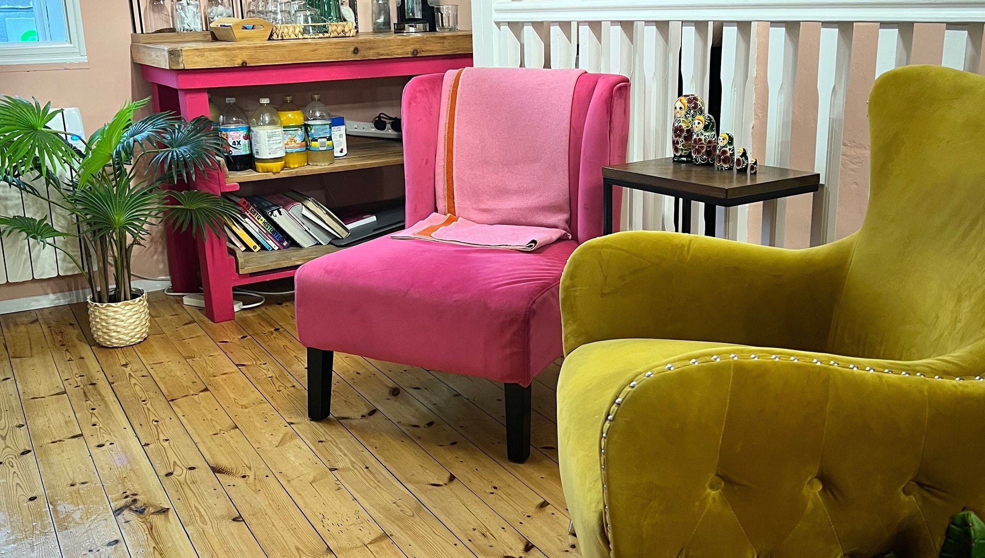 Inside the salon: a hot pink velvet chair and mustard velvet armchair on warm pine floorboards, with plants and a pink painted drinks console